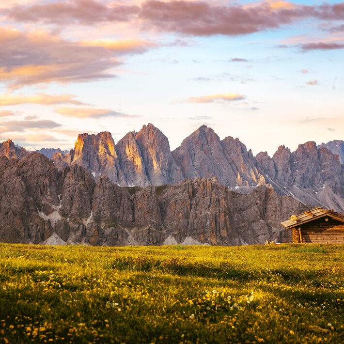 Mountain meadow with a small wooden hut and Dolomite peaks at sunrise. | © Matthias Gasser