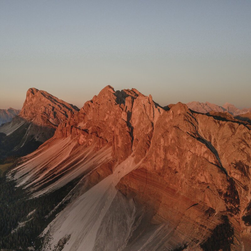 Cime dolomitiche illuminate al tramonto con creste rocciose ripide e pendii boscosi sotto.