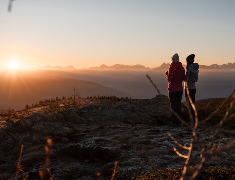 Zwei Wanderer auf einem Felsgrat bei Sonnenaufgang mit Blick auf ferne Gipfel. | © Brandnamic