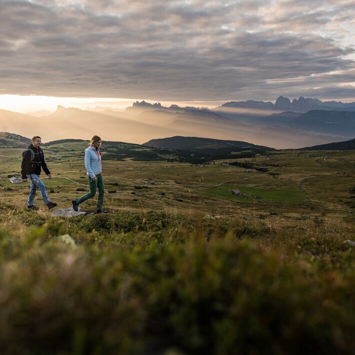 Two hikers walking across an alpine plateau at sunrise with mountain views. | © (c) Harald Wisthaler