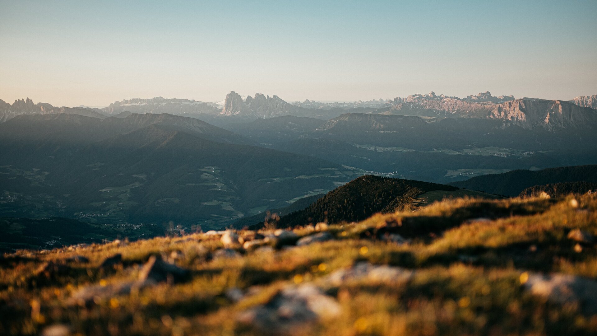 Sunlit alpine meadow with distant mountain peaks and a wide valley under a clear sky. | © (c) Tobias Kaser