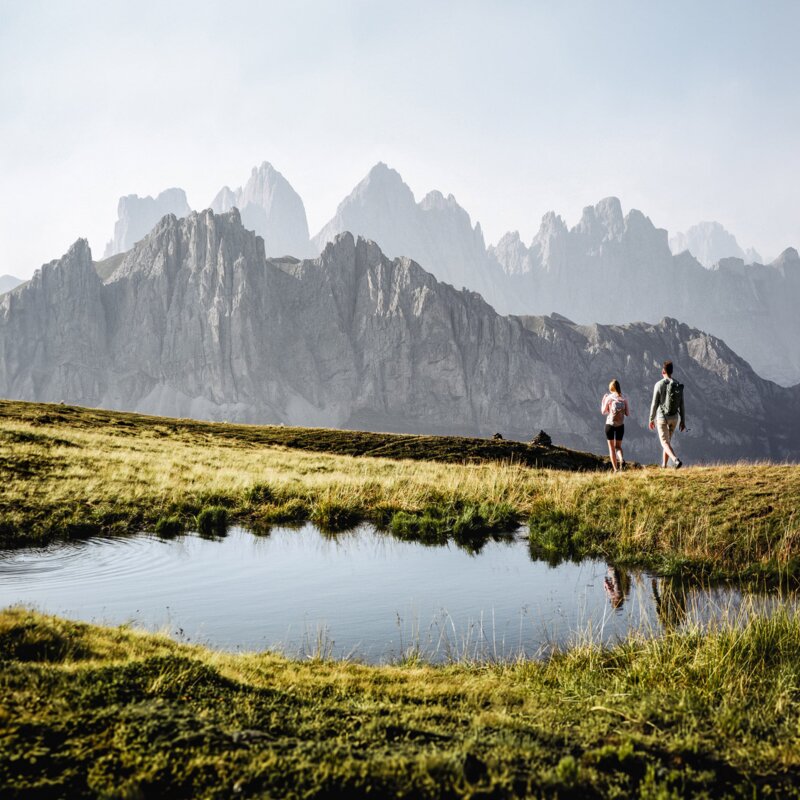 Hikers on a grassy alpine plateau beside a small mountain pond with jagged peaks behind. | © (c) Argento-Artistry