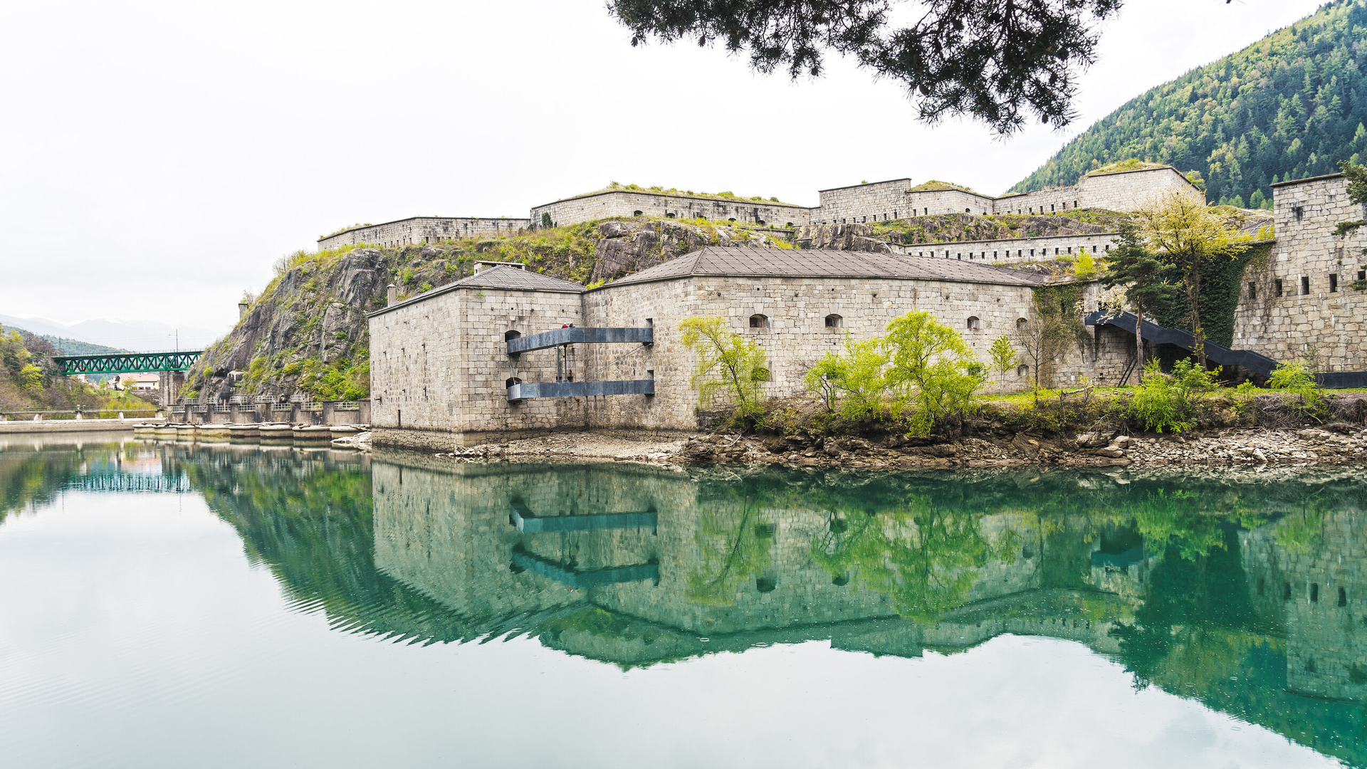 Stone fortress reflected in calm green water beneath mountain slopes