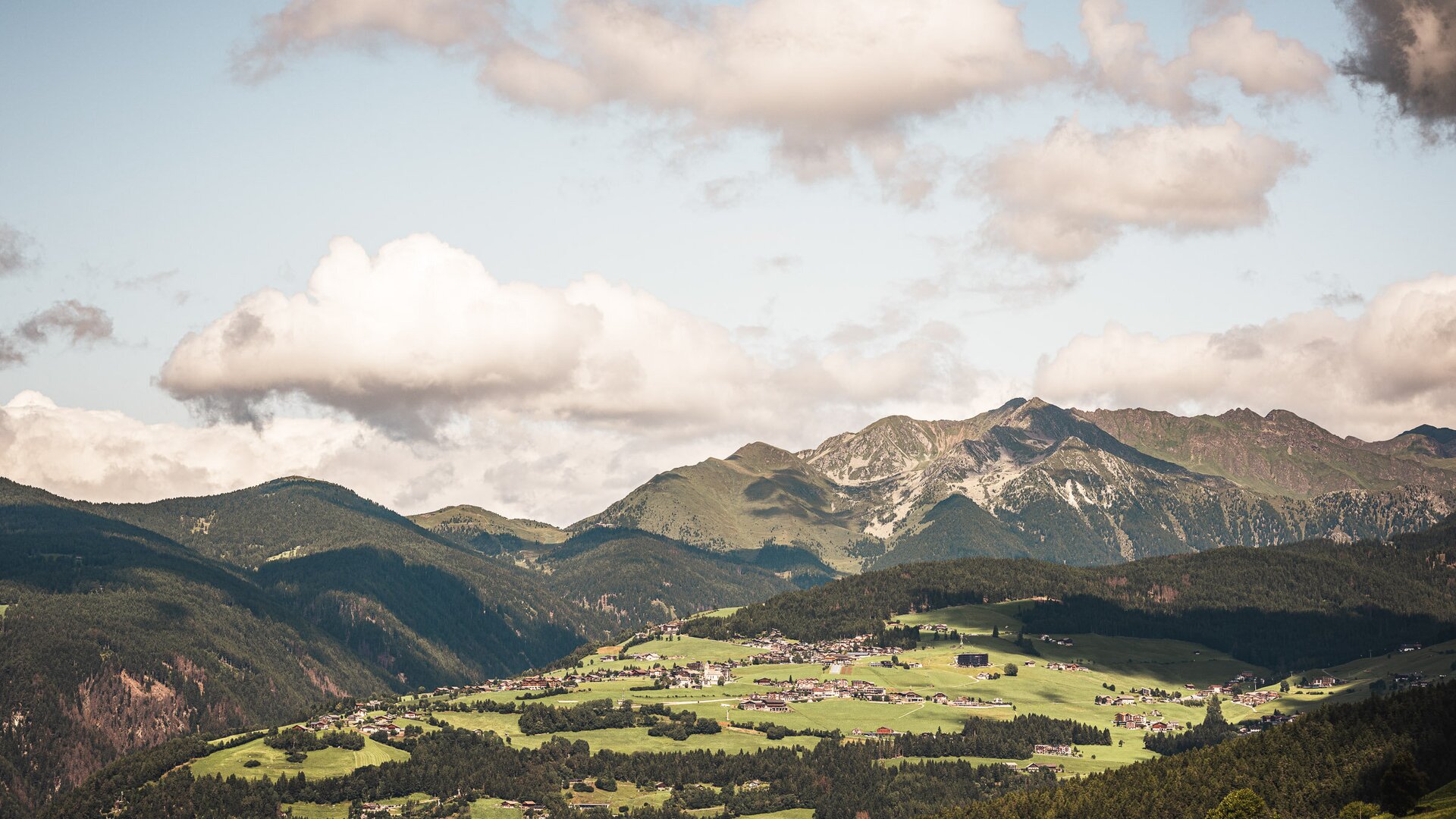 Green alpine valley with scattered villages and mountain peaks under a cloudy sky. | © (c) Hannes Niederkofler