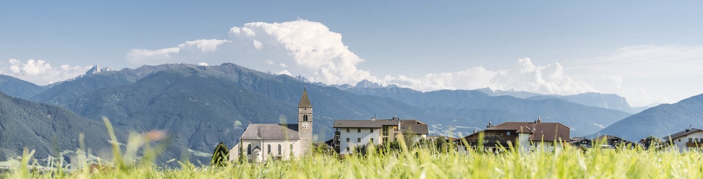 Kirche und Häuser eines Dorfes hinter einer grünen Wiese mit Bergkulisse in Brixen Südtirol.