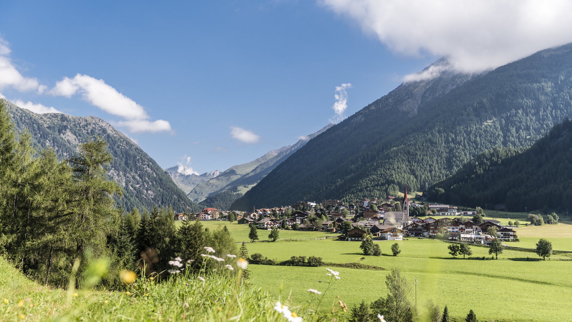 Sunny valley with village, green meadows and mountain slopes under a blue sky.