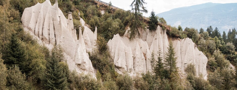 Piramidi di terra bianche su un pendio verde con alberi e montagne sullo sfondo. | © (c) Brandnamic