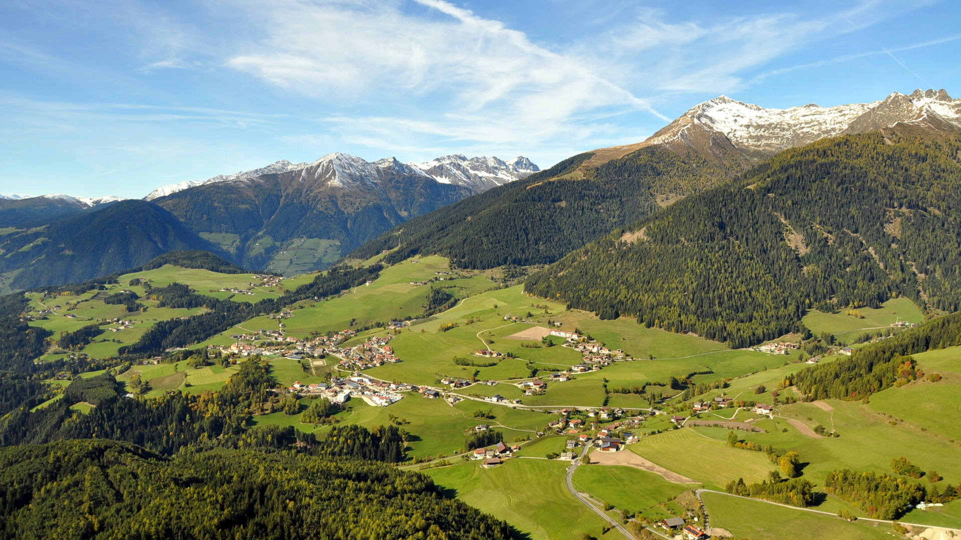 Aerial view of a green alpine valley with villages, forested slopes and snow-capped mountains.