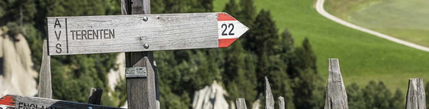 Holzwegweiser bei Terenten über grünen Almwiesen und bewaldeten Hängen.