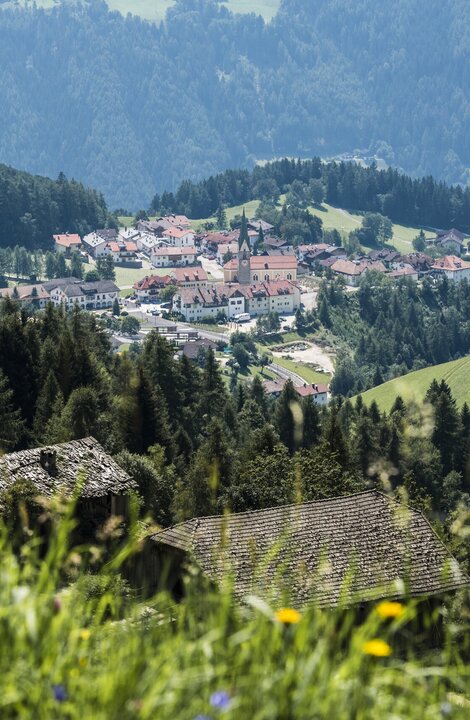 Blick auf ein Taldorf mit Kirche, Wiesen und bewaldeten Hängen im Sommer.