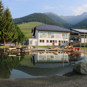 Teich mit Spiegelung von Gebäude, Bäumen und Bergen in einem sonnigen Alpental.