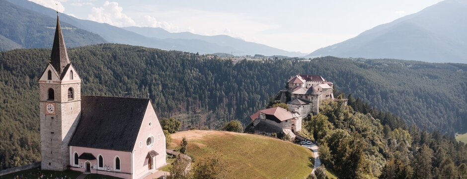 Kirche auf einem Hügel mit Friedhof, Burgruine und bewaldeten Bergen im Hintergrund.