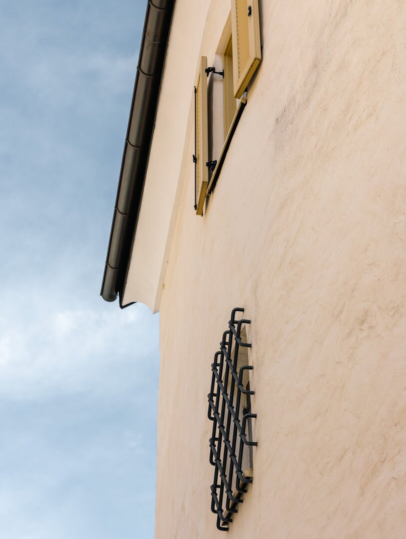Cremefarbene Hausfassade mit Fensterladen und Wandgitter vor blauem Himmel