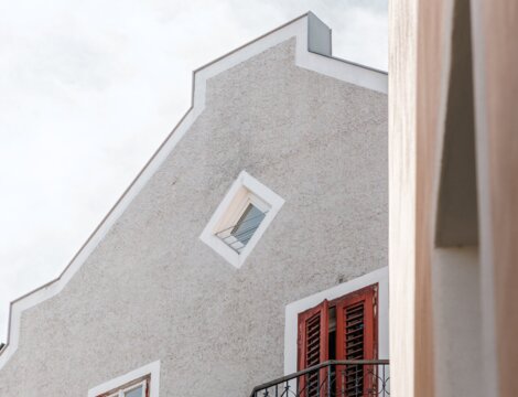 Graue Hausfassade mit roten Fensterläden, Balkon und hellem Himmel darüber