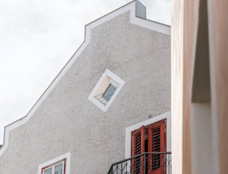 Graue Hausfassade mit roten Fensterläden, Balkon und hellem Himmel darüber