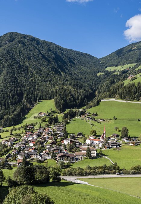 Grünes Alpental mit Dorf, Kirchturm und bewaldeten Bergen unter blauem Himmel.
