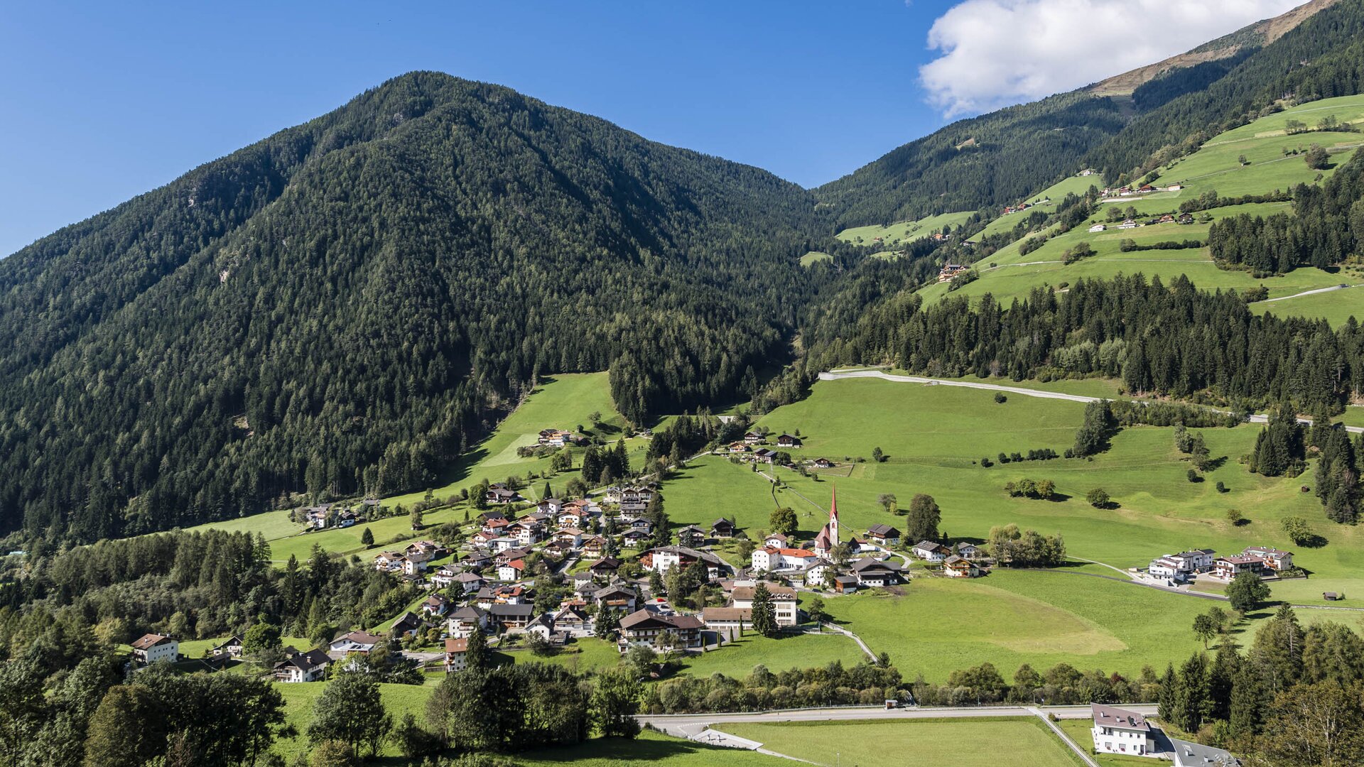 Green alpine valley with a village, church steeple and forested mountains under a blue sky.