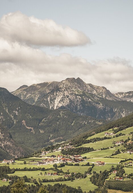 Grünes Bergtal mit kleinem Dorf und felsigen Gipfeln unter bewölktem Himmel.