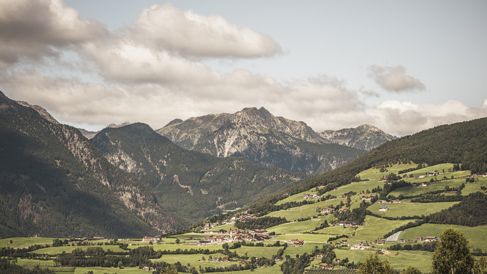 Green mountain valley with a small village and rocky peaks under cloudy skies.