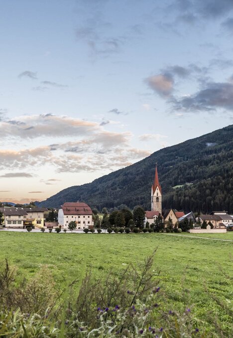 Dorf im grünen Tal mit Kirche, umgeben von bewaldeten Bergen unter einem sanften Abendhimmel.