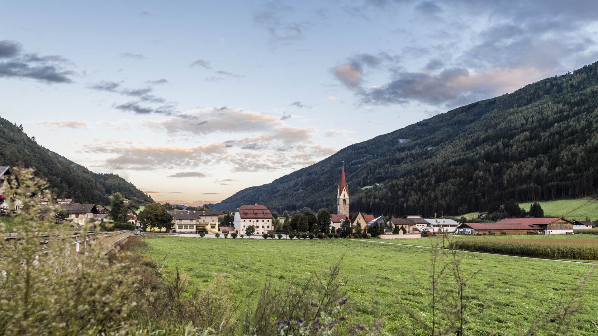 Village in a green valley with a church, surrounded by forested mountains under a soft evening sky.