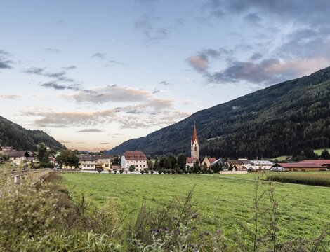 Paese in una valle verde con una chiesa, circondato da montagne boscose sotto un cielo serale delicato.