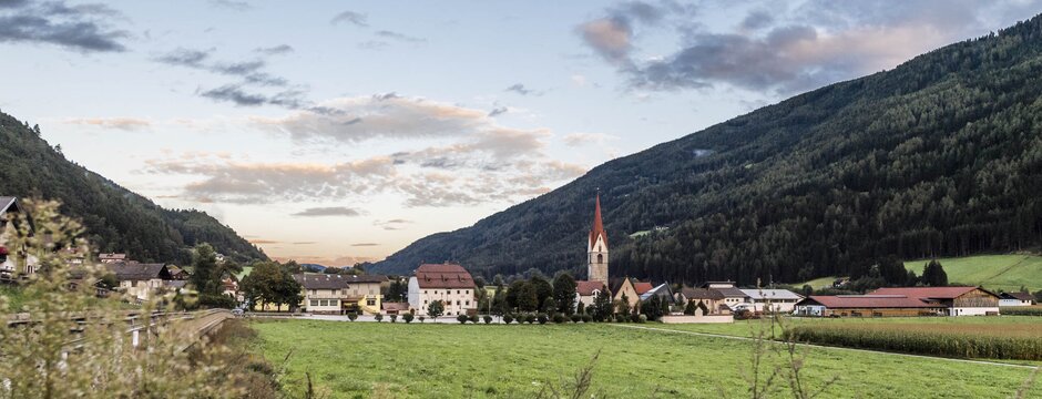 Dorf im grünen Tal mit Kirche, umgeben von bewaldeten Bergen unter einem sanften Abendhimmel.