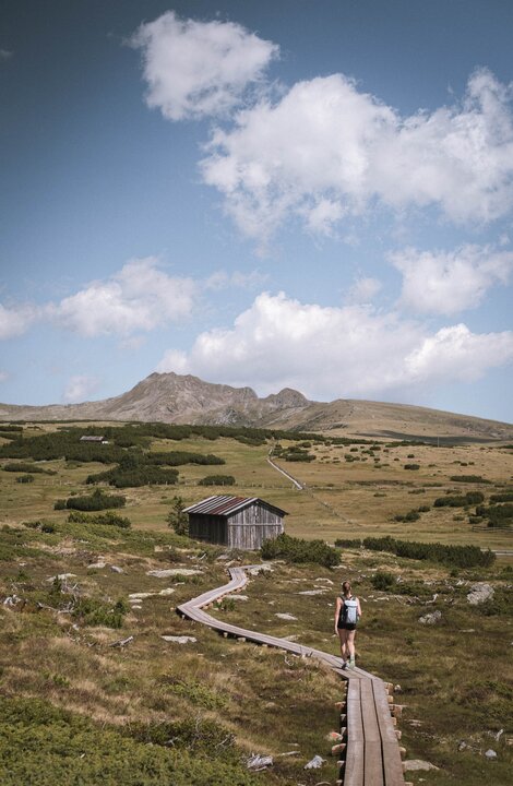 Wanderin auf einem Holzsteg über ein alpines Plateau mit Hütte und Berggipfeln unter blauem Himmel.