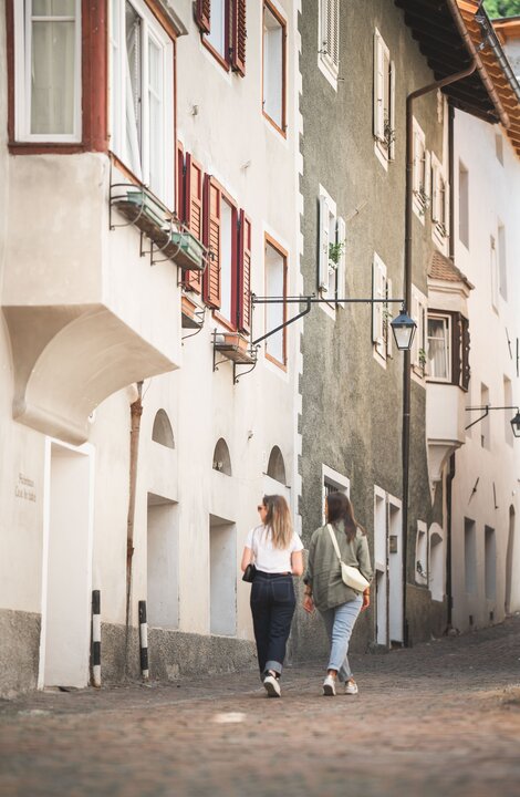 Two women walking through a narrow cobblestone street in Brixen Südtirol's old town. | © Koni Studios