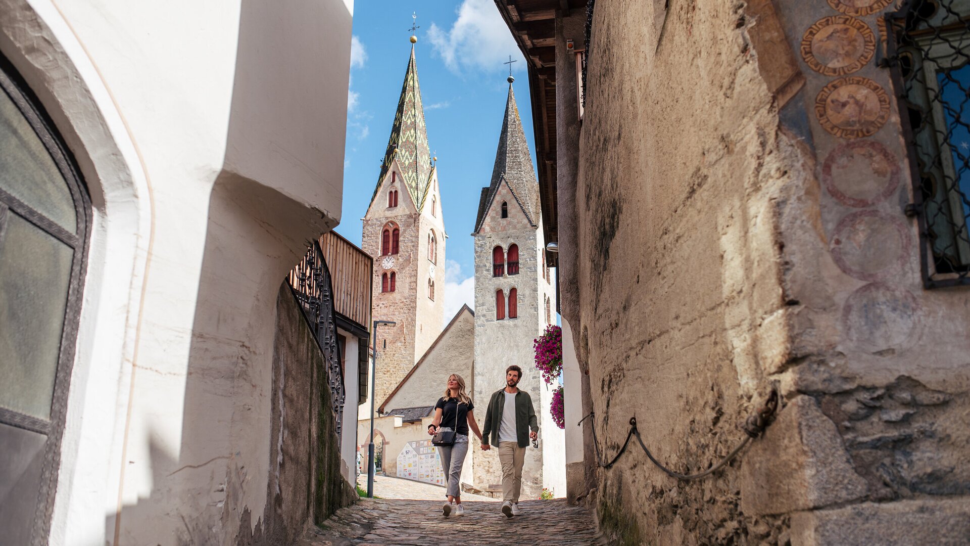Couple walking through a cobblestone alley in Brixen Südtirol with church towers and blue sky
