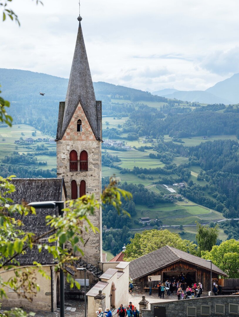 Church tower above a village with a wide green valley and mountains in the background.