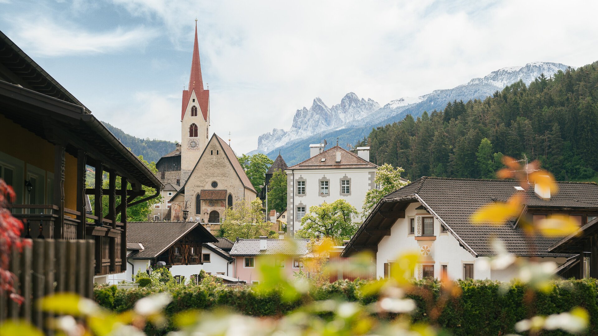 Village church with a red spire in Brixen Südtirol, framed by alpine houses and mountain peaks.