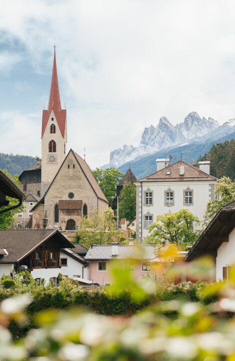 Village church with a red spire in Brixen Südtirol, framed by alpine houses and mountain peaks.