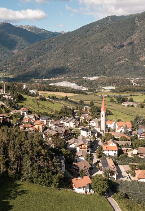 Borgo con campanile in una valle verde sotto le montagne.