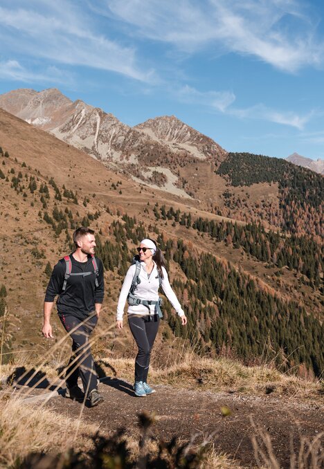 Due escursionisti su un sentiero di montagna con cime alpine e una valle boscosa sullo sfondo.