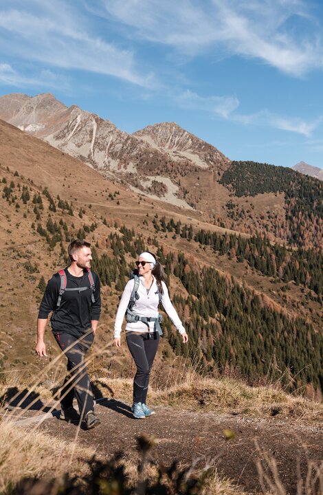 Two hikers on a mountain trail with alpine peaks and a forested valley in the background.