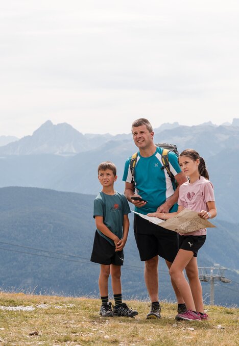 Famiglia con due bambini in cammino su un plateau di montagna con cartina e panorama alpino