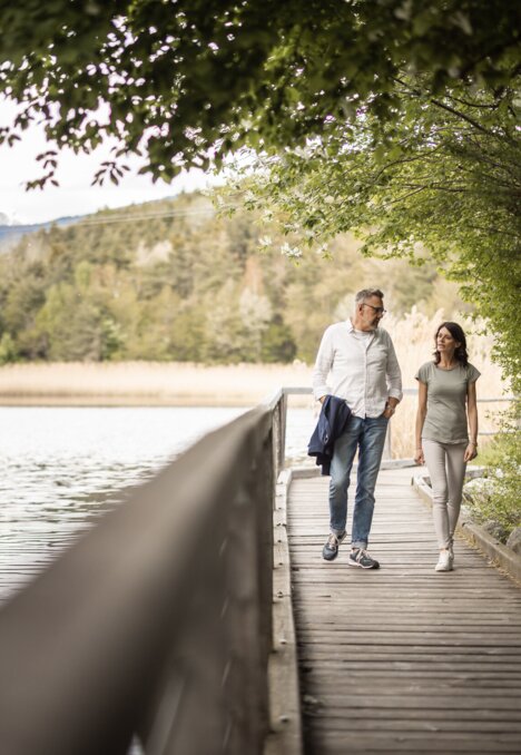 Couple walking along a lakeside boardwalk with mountains and trees in the background.