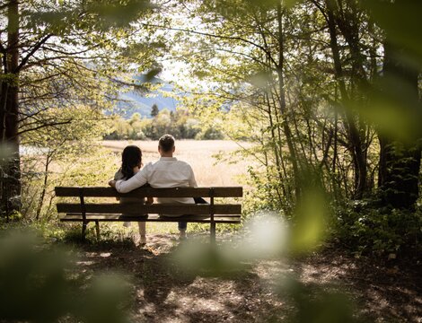 Couple sitting on a bench in a shaded forest, looking out over a sunny meadow and distant mountains.