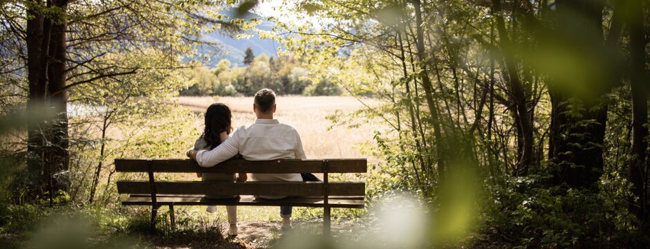 Couple sitting on a bench in a shaded forest, looking out over a sunny meadow and distant mountains.
