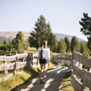 Woman walking along a fenced path through a sunny meadow with trees and mountains in the background.