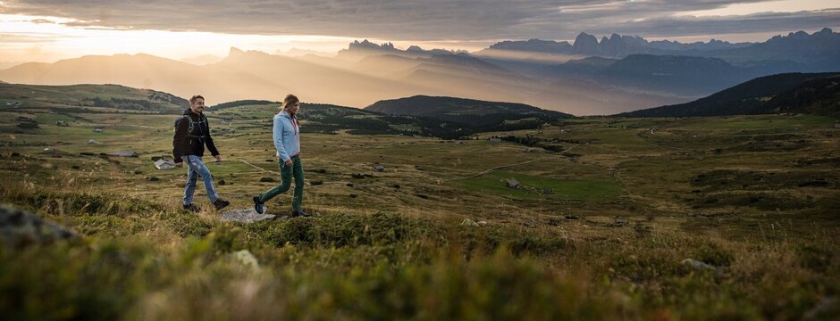 Two hikers walking across a grassy plateau with mountain peaks in the distance at sunrise near Brixen Südtirol.