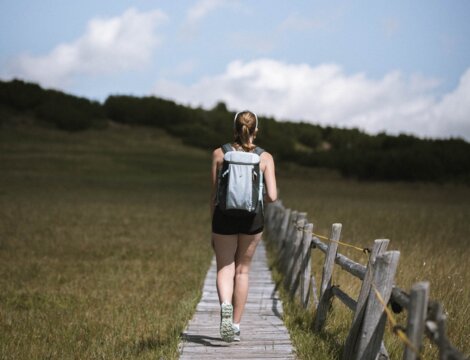 Woman hiking on a wooden path through a green meadow under a blue sky.