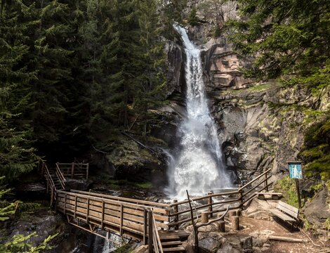 Holzsteg mit Treppe zu einem Wasserfall in einer bewaldeten Schlucht.