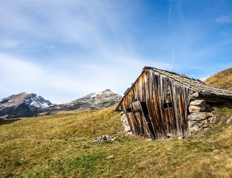 Vecchio rifugio alpino in un prato con cime innevate e cielo azzurro