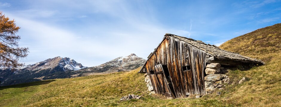 Old alpine hut in a grassy meadow with snow-dusted mountains and blue sky