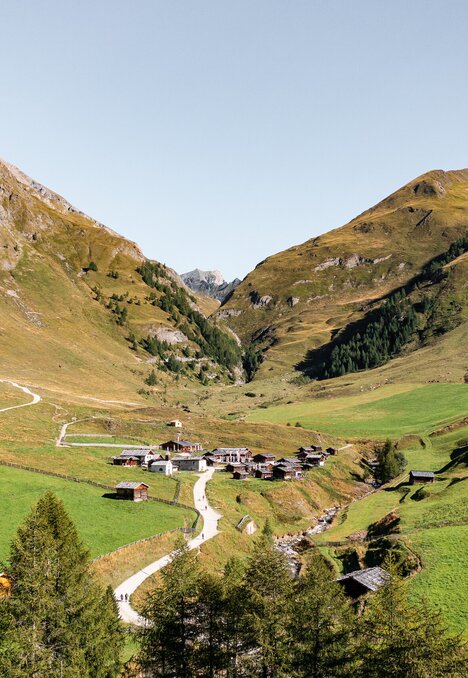 Grünes Alpental mit kleinem Dorf und Bergflanken unter klarem Himmel.