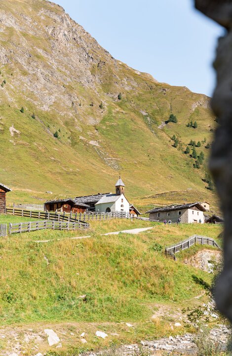 Kleines Alpendorf mit Kirche in einem grünen Talkessel unter einem steilen Felshang.