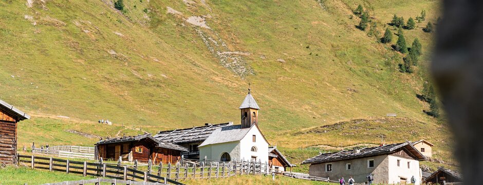 Piccolo borgo alpino con chiesa in una valle verde sotto un ripido pendio roccioso.