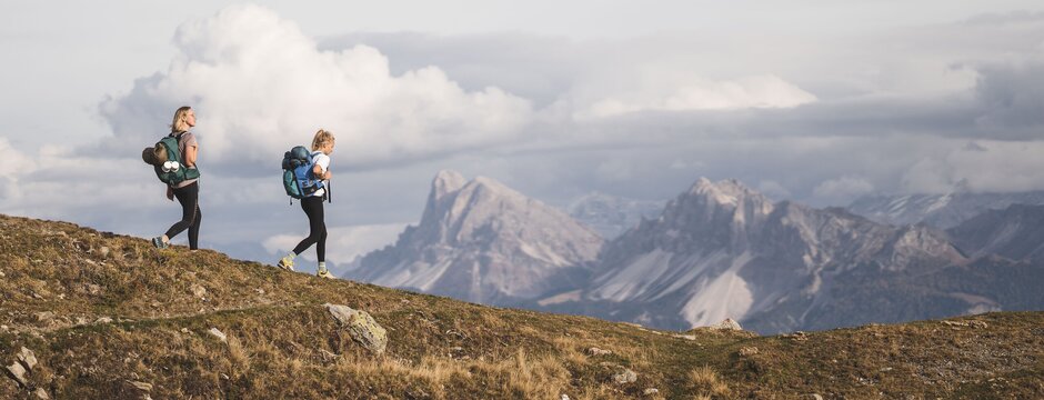 Zwei Wanderer gehen auf einem Berggrat mit Gipfeln und Wolken im Hintergrund.